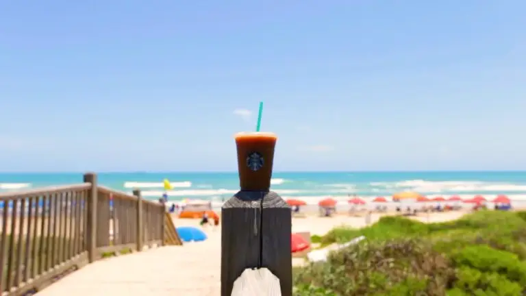 A coffee on the boardwalk rail with umbrellas on the beach behind it